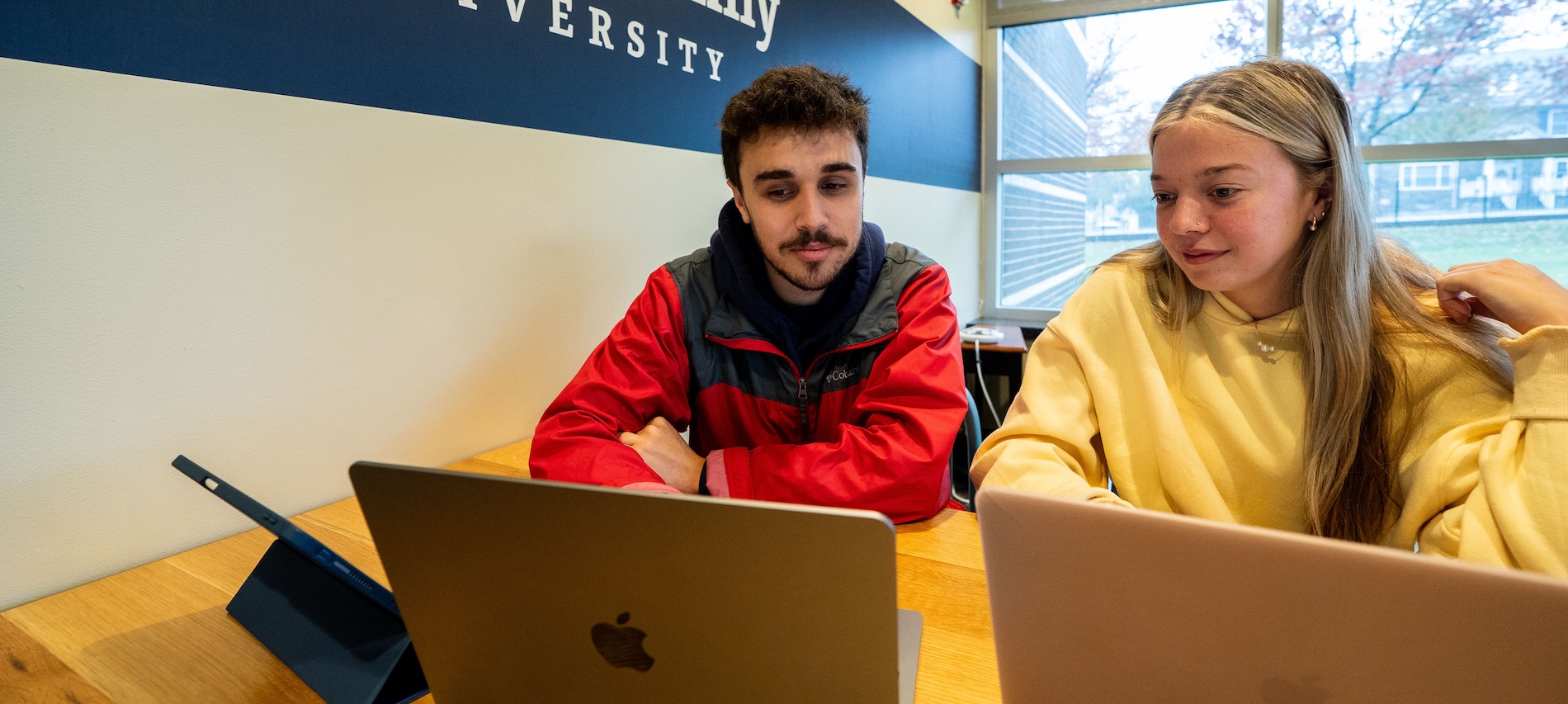 Two students with laptops helping each other study in a breakout room on campus