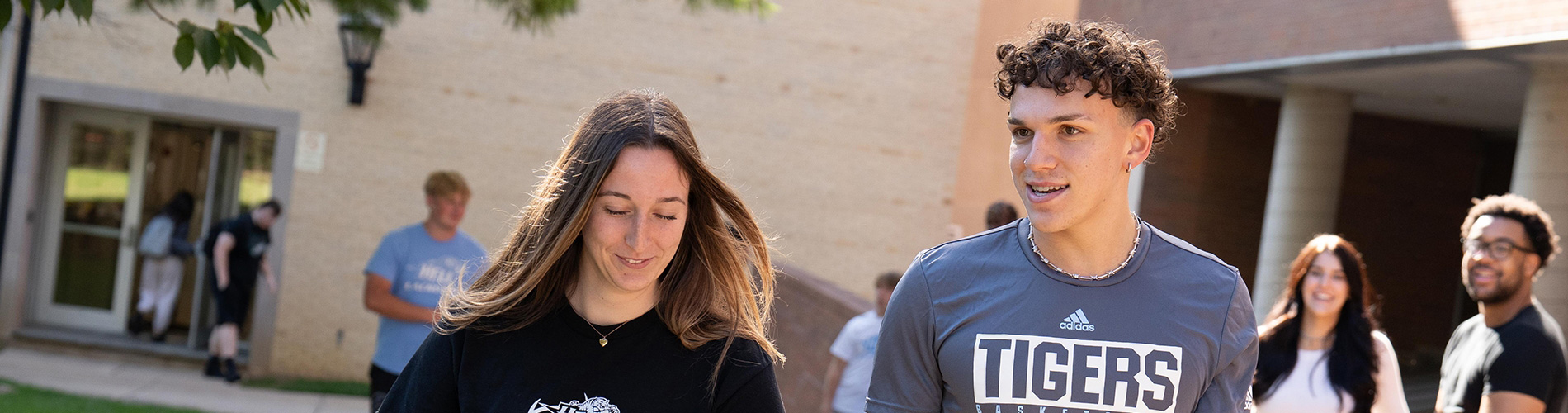 Two students walking and talking on a path through campus while other students socialize in the background
