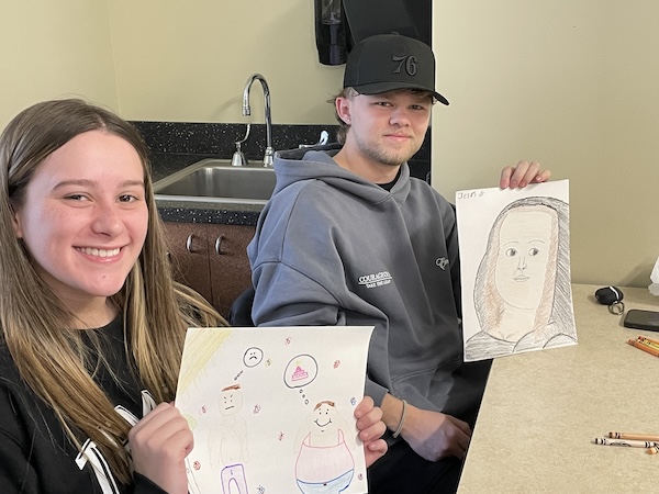 Two students at their desks in the classroom holding up their artwork