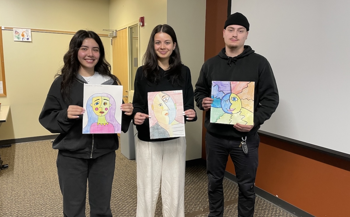 Three HFU students holding up their art projects at the front of a classroom