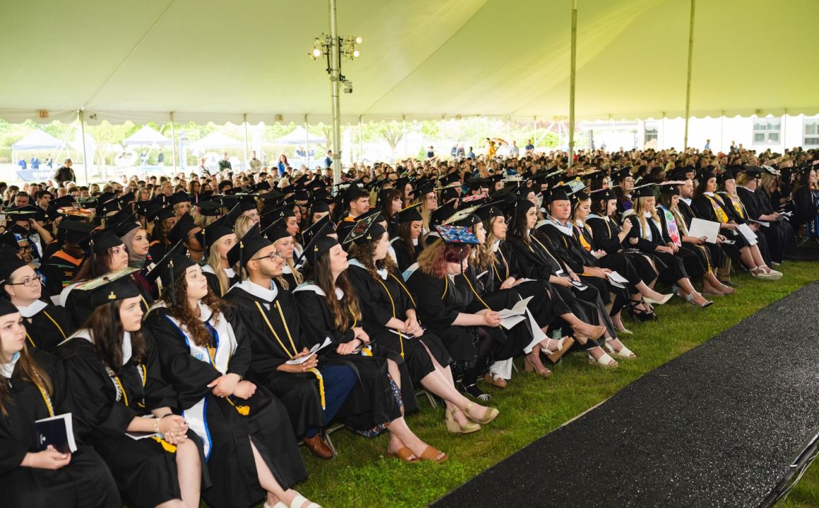 Large crowd of HFU students in caps and gowns sitting under a text during 2024's Commencement